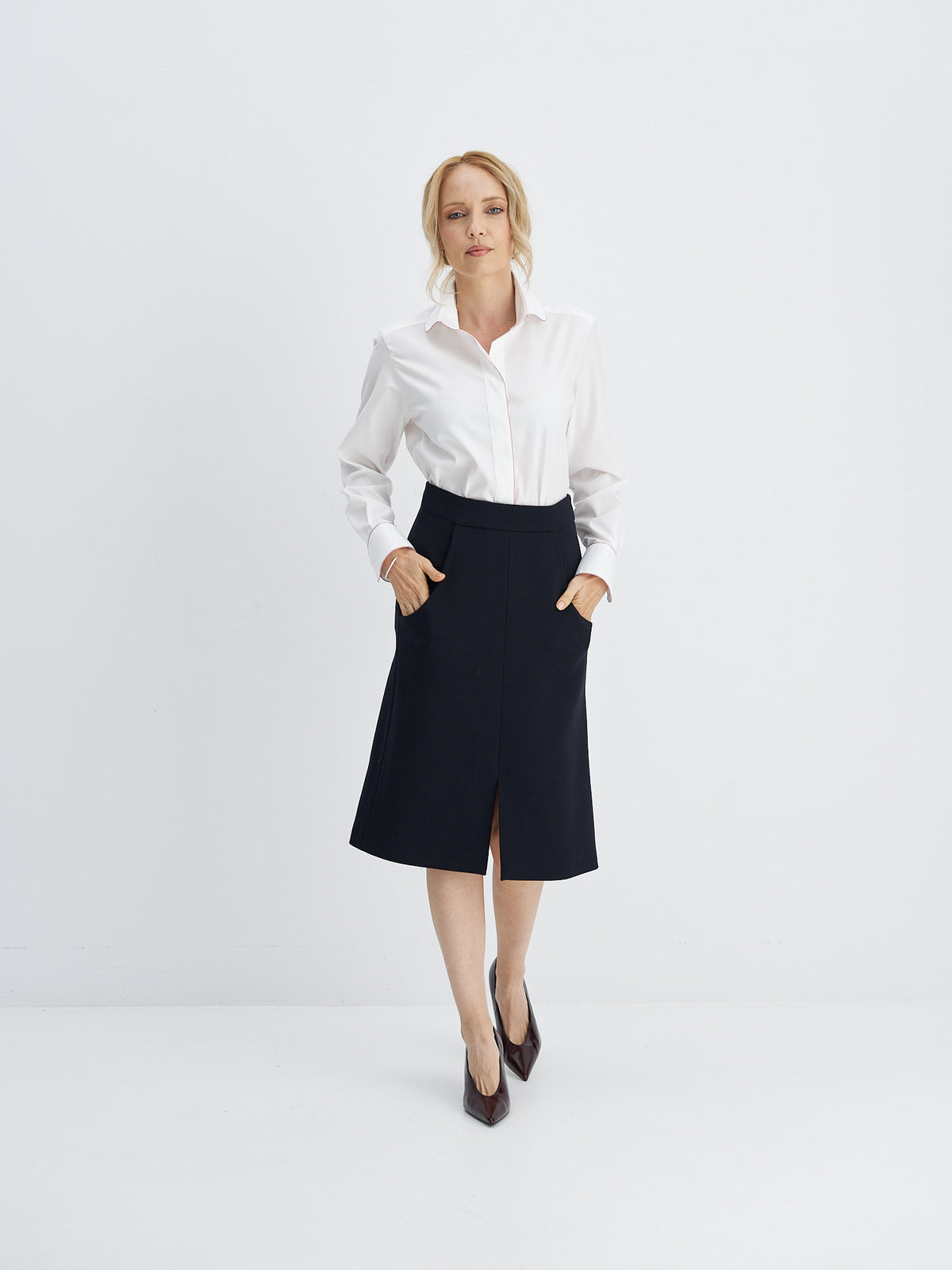 A woman modeling a white long-sleeve button-down shirt and a dark navy knee-length skirt with front pockets, paired with dark brown pointed-toe heels, against a clean white studio background.