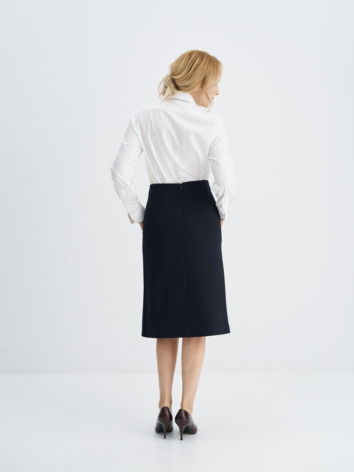 A woman is shown from behind wearing a white long-sleeve button-up shirt and a dark navy knee-length skirt, paired with dark brown pointed-toe heels, against a clean white studio background.