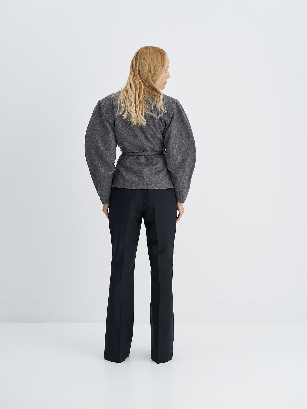 Back view of charcoal grey belted wool jacket showing curved sleeves and a defined waist, worn with black trousers