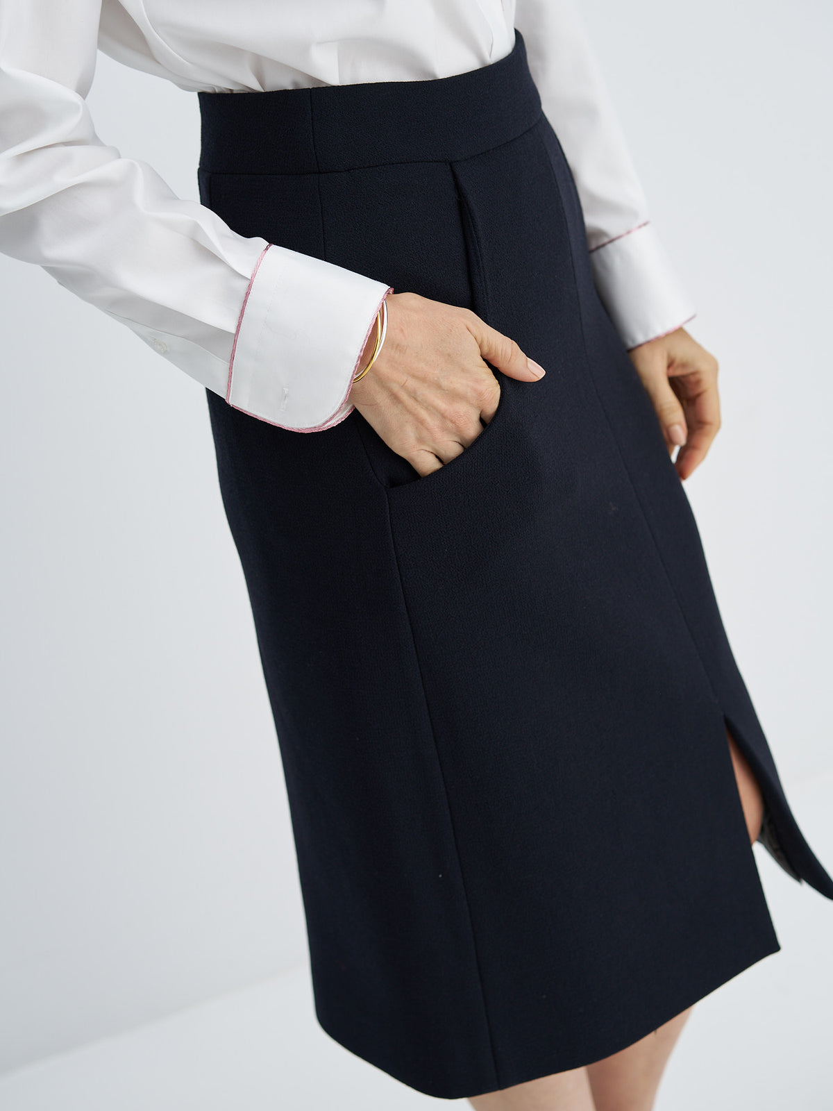 A close-up of a model wearing a dark navy blue skirt with a side slit and pocket, paired with a white long-sleeve shirt with pink piping on the cuffs, against a light gray background.