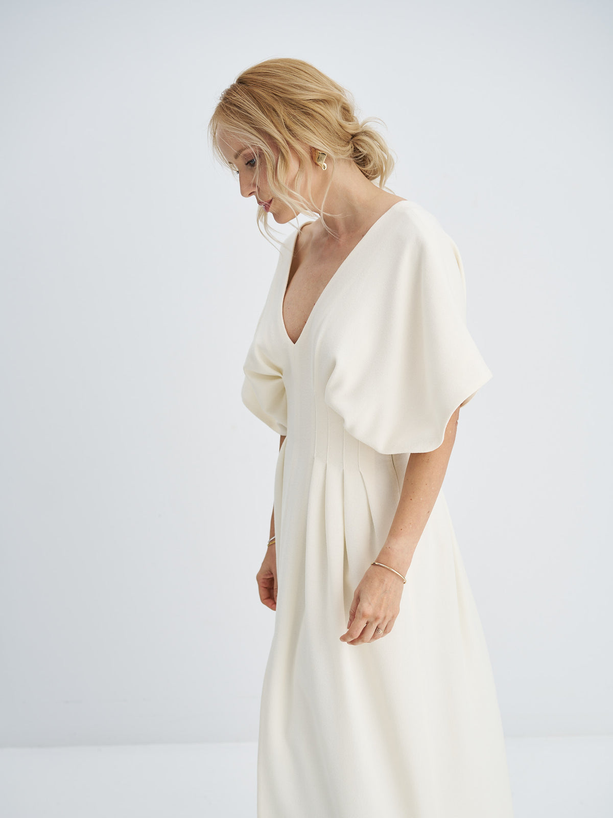 Three-quarter view of ivory wool-crepe dress showing the draped sleeve and fitted pleated waist