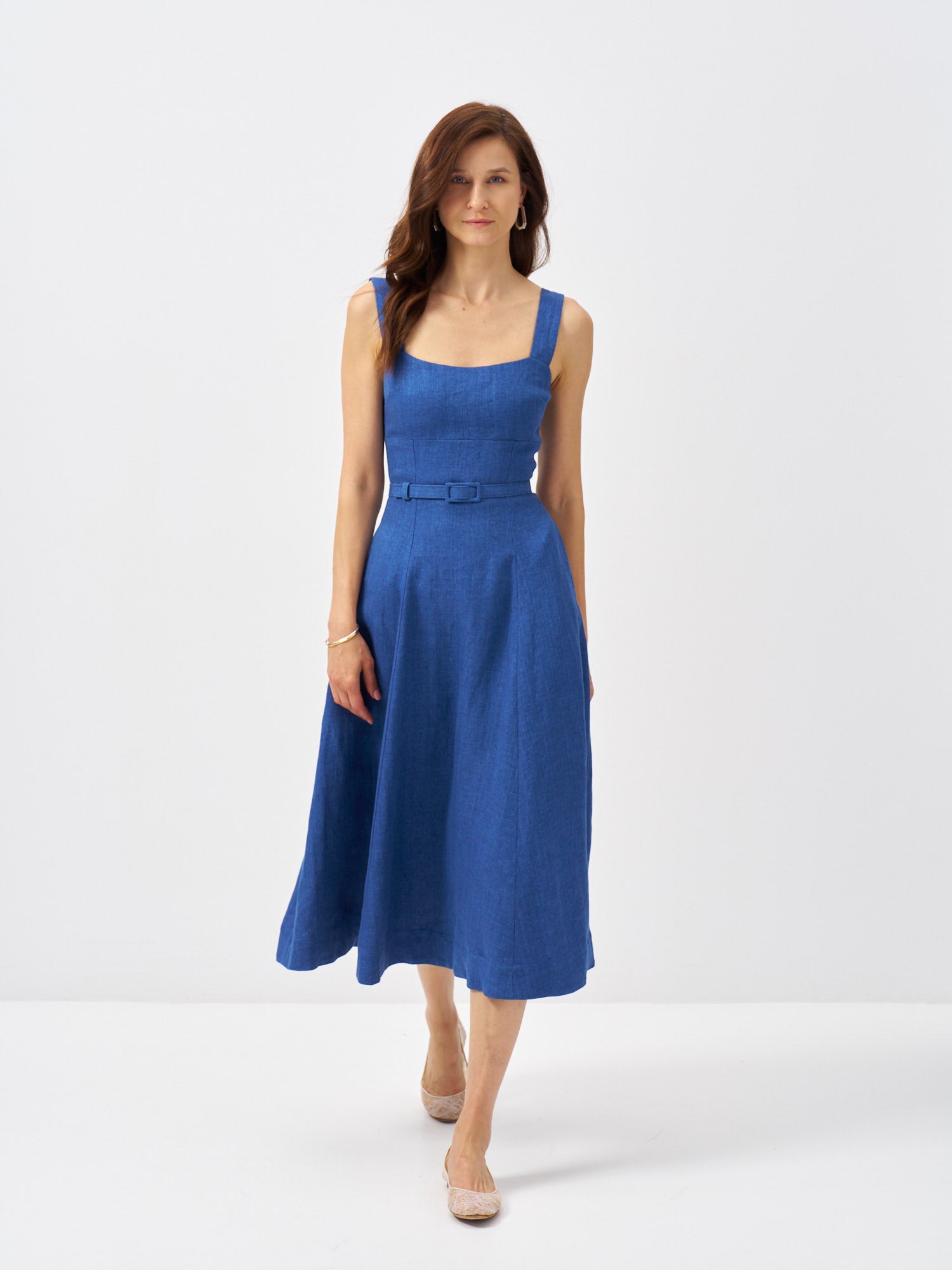 Woman wearing a blue linen midi dress with wide straps, fitted bodice, matching belt and full skirt.